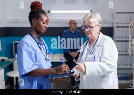 Une infirmière afro-américaine signe des papiers de contrôle alors que le médecin tient la planchette à pince dans le cabinet.Assistant médical et médecin examinant les informations sur les soins de santé et les données sur les dossiers lors de la visite de contrôle Banque D'Images