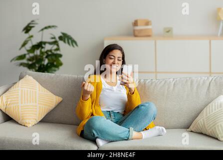 Oui.Femme arabe excitée utilisant un gadget de smartphone, se serrer le poing, célébrer la victoire, porter des écouteurs sans fil Banque D'Images