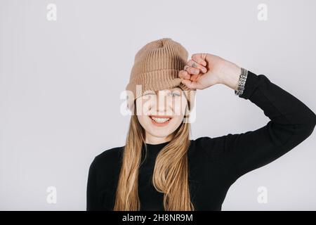 Portrait affectif franc et non modifié de jeune blonde heureux cheveux longs femme couvrant les yeux avec bonnet beanie tricoté marron.Portrait de sourire amusant Banque D'Images