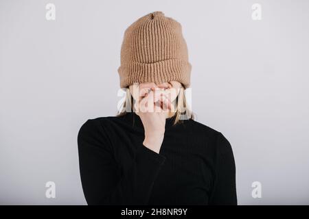 Portrait affectif franc et non modifié de jeune blonde heureux cheveux longs femme couvrant les yeux avec bonnet beanie tricoté marron.Portrait de sourire amusant Banque D'Images