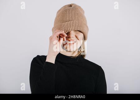 Portrait affectif franc et non modifié de jeune blonde heureux cheveux longs femme couvrant les yeux avec bonnet beanie tricoté marron.Portrait de sourire amusant Banque D'Images