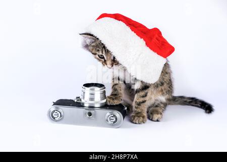 Petit chaton mignon avec chapeau de Père Noël et appareil photo vintage sur fond blanc. Banque D'Images