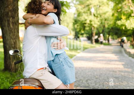 Couple multiethnique souriant qui s'embrasse assis sur une moto dans le parc Banque D'Images