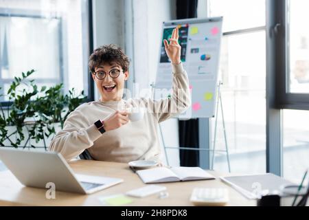 Homme d'affaires enjoué avec une tasse ayant des appels vidéo sur ordinateur portable près de l'ordinateur portable dans le bureau, image de stock Banque D'Images