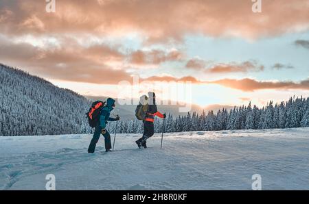 Deux randonneurs vêtus de vêtements de sport d'hiver chauds avec des sacs à dos de randonnée marchent avec des bâtons de randonnée dans les montagnes de pins enneigées dans un incroyable coucher de soleil Banque D'Images