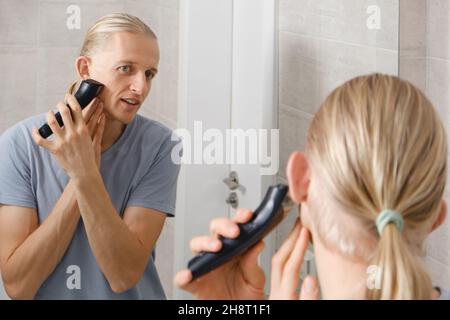 L'homme qui se rasante murmure les cheveux lui-même, coupe-cheveux à la maison devant le miroir dans le bain.Homme avec une coupe longue à l'aide de la tondeuse pour machine à rasage Banque D'Images