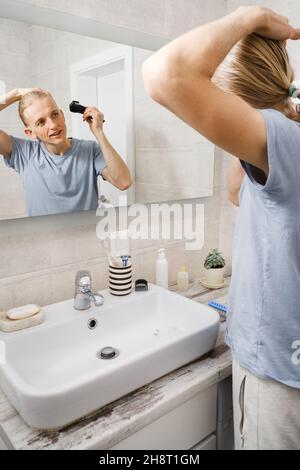 L'homme qui se rasante murmure les cheveux lui-même, coupe-cheveux à la maison devant le miroir dans le bain.Homme avec une coupe longue à l'aide de la tondeuse pour machine à rasage Banque D'Images