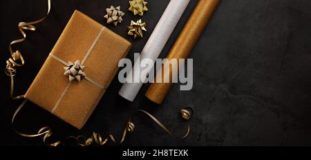 service d'emballage de cadeaux. boîte cadeau dorée emballée avec rouleaux de papier d'emballage sur fond de pierre sombre. espace de copie de bannière Banque D'Images