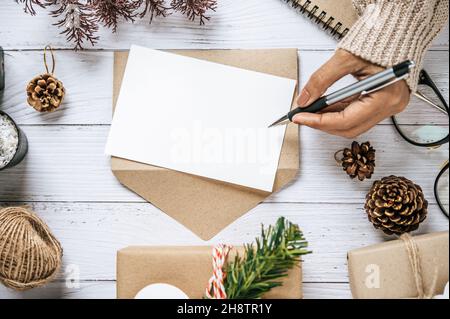 Une main tenant un stylo pour écrire sur un presse-papiers a été placée sur une table en bois blanc avec des verres, un stylo et des cônes de pin séchés. Banque D'Images