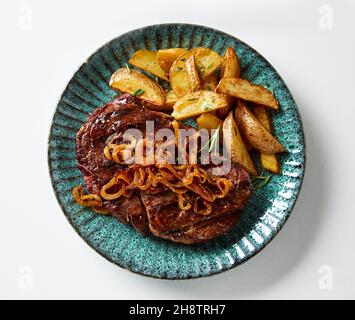 Vue de dessus du steak de bœuf grillé avec oignon frit et coin de pommes de terre rustique servis sur l'assiette sur une table blanche Banque D'Images