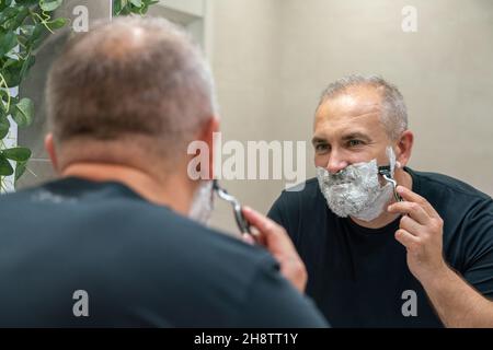 Homme mûr aux cheveux gris recoiffant lui-même sa barbe à la maison à l'aide d'un rasoir Banque D'Images