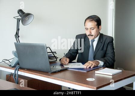 Cadre d'affaires d'âge moyen avec moustache portant un costume et une cravate écrivant avec un stylo sur son poste de travail de bureau. Banque D'Images