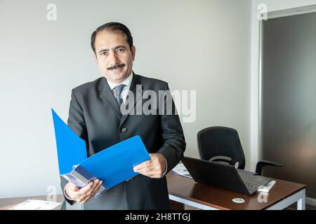 Cadre d'affaires d'âge moyen avec moustache portant un costume et une cravate tenant une chemise bleue sur son poste de travail de bureau. Banque D'Images