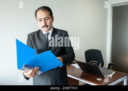 Cadre d'affaires d'âge moyen avec moustache en costume et cravate lisant le dossier bleu sur son poste de travail de bureau. Banque D'Images