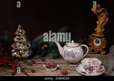 Théière et tasse de thé de Noël, bonbons dans une assiette en céramique, branches d'arbre de Noël, arbre de Noël en céramique et une horloge antique sur une table en bois. Banque D'Images