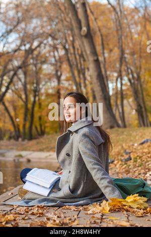 Jeune belle écolière assise dans un parc d'automne avec un manuel Banque D'Images