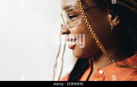 Jeune femme souriant gaiement en se tenant seule à l'intérieur.Vue latérale d'une jeune femme heureuse portant des lunettes et des braïdes. Banque D'Images