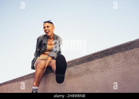 Jeune femme gaie s'amusant en s'asseyant sur un mur à l'extérieur.Une jeune femme heureuse rit et se fait un bon moment.Jeune femme hipster sentiment Banque D'Images