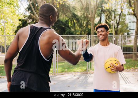 Jeunes athlètes de race masculine de dos avec des mains de basket-ball jaunes tout en se tenant debout sur le terrain de sport dans le parc regardant l'un l'autre Banque D'Images