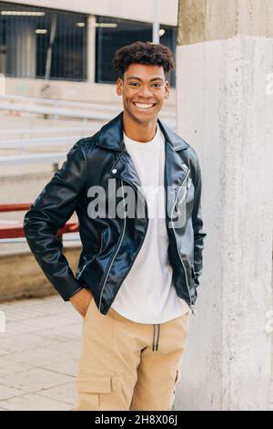 Jeune homme noir gai dans une veste en cuir debout dans la rue avec les mains dans les poches près de la colonne et regardant l'appareil photo Banque D'Images