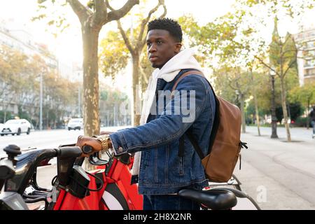 Vue latérale d'un homme afro-américain avec des cheveux fclés foncés dans une veste en denim debout et en train de prendre un vélo à la station de location dans la rue à la lumière du jour Banque D'Images