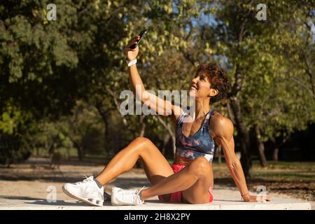 Corps complet de la bonne ethnie adulte Sportswoman avec des cheveux courts et bouclés dans les vêtements d'activité et les baskets souriant et prenant le selfie sur le smartphone après un traa extérieur Banque D'Images