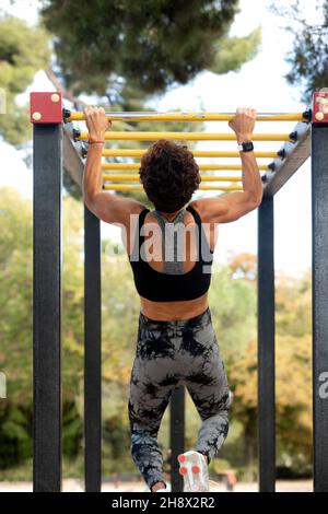 Vue arrière d'une Sportwoman méconnaissable en casque d'activité suspendu sur un bar en métal lors d'un entraînement sur un terrain de sport dans le parc en journée Banque D'Images
