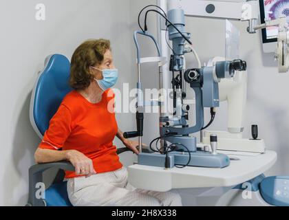 Vue latérale d'une patiente senior sous masque médical de protection, assise sur un équipement ophtalmologique d'ophtalmoscope Banque D'Images