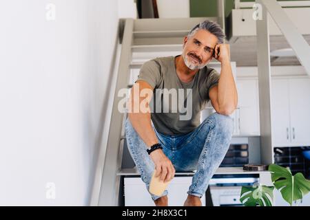 Homme âgé gai pieds nus assis sur une échelle et regardant la caméra tout en se penchant à la main dans un appartement lumineux Banque D'Images