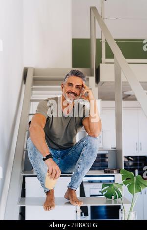 Homme âgé gai pieds nus assis sur une échelle et regardant la caméra tout en se penchant à la main dans un appartement lumineux Banque D'Images