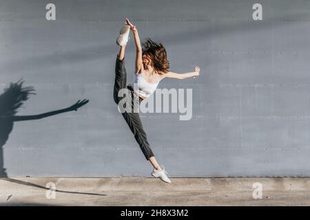 Corps complet de gymnaste féminin anonyme flexible en vêtements de sport touchant la jambe fendue tout en sautant contre le mur gris Banque D'Images