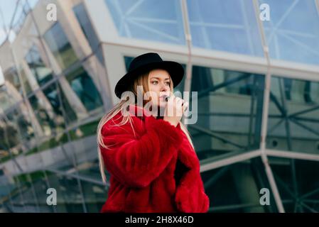 Jolie jeune femme blonde avec chapeau et veste rouge fumage avec machine à couches dans la rue Banque D'Images
