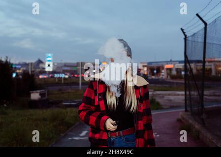 Jolie jeune femme blonde avec chapeau de laine debout sur la rue et fumer avec machine à couches Banque D'Images