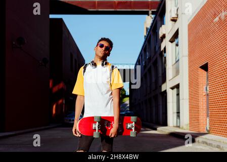 Jeune homme noir avec lunettes de soleil tenant un skateboard posé dans la rue Banque D'Images
