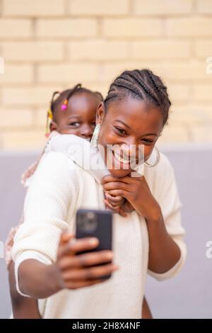 Jeune mère noire gaie avec des tresses afro dans des vêtements décontractés souriant et utilisant un smartphone tout en prenant selfie avec adorable petite fille à l'arrière Banque D'Images