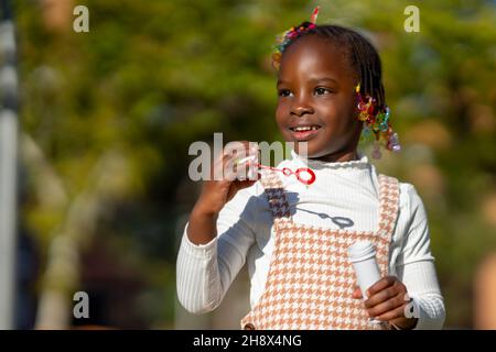 Adorable petite fille afro-américaine heureuse avec des cheveux colorés sur les tresses souriant et regardant loin tout en soufflant des bulles de savon dans le parc vert sur su Banque D'Images