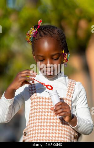 Adorable petite fille afro-américaine heureuse avec des cheveux colorés sur des tresses soufflant des bulles de savon dans le parc vert le jour ensoleillé Banque D'Images