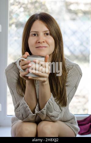 Femme assise sur le rebord de la fenêtre avec une tasse de thé chaud Banque D'Images