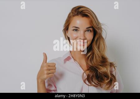 Jeune fille attrayante avec des cheveux magnifiquement stylisés montrant le pouce vers le haut, debout isolé sur gris Banque D'Images