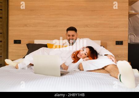 Un jeune couple de peignoirs est allongé sur le lit avec un ordinateur portable.Homme et femme regardant un film.Une femme repose avec sa tête sur les genoux d'un homme. Banque D'Images