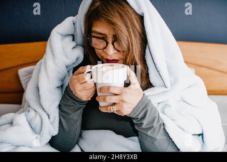 Femme hispanique couverte de couvertures buvant une tasse de chocolat chaud avec des guimauves Banque D'Images