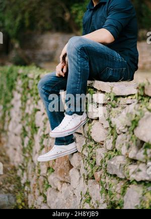 Homme en jeans et baskets assis sur un mur de pierre dans le parc.Gros plan Banque D'Images