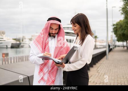 Homme arabe chantant contrat de charte afin de réserver yacht, homme d'affaires saoudien en port traditionnel faisant affaire avec une jeune femme caucasienne courtier tout en se tenant ensemble dans le port Banque D'Images