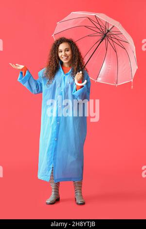 Jeune femme afro-américaine portant un imperméable bleu et des bottes de costume avec parapluie sur fond rouge Banque D'Images