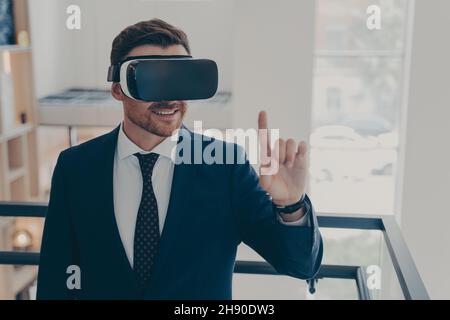 Homme d'affaires souriant et réussi en costume debout à l'intérieur du bureau et utilisant des lunettes VR pour le travail Banque D'Images