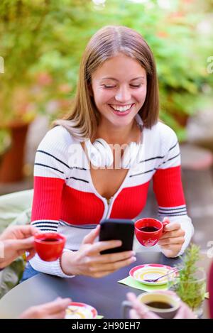 Une jeune femme heureuse souriant avec plaisir car elle utilise son smartphone tout en se détendant à une table en prenant une tasse de café avec des amis dans un restaurant en plein air Banque D'Images