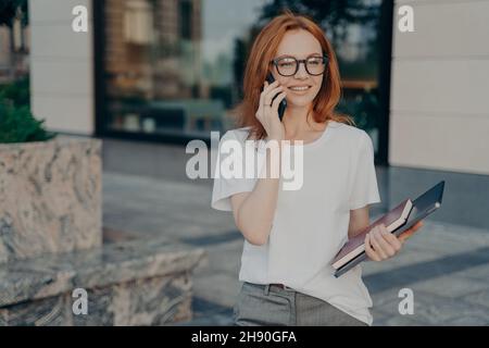 Jeune femme d'affaires souriante dans des lunettes parlant sur un téléphone mobile à l'extérieur Banque D'Images