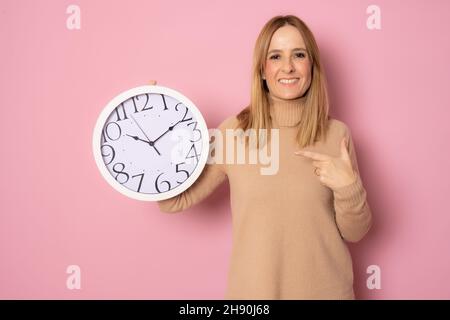 Bonne femme caucasienne pointant du doigt vers la grande horloge isolée sur fond rose. Banque D'Images