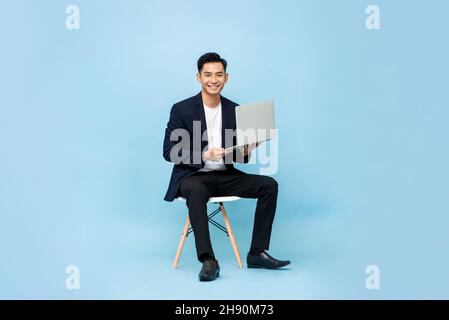 Portrait d'un jeune homme d'affaires souriant et charmant d'Asie du Sud-est avec ordinateur portable assis sur une chaise sur fond de studio bleu clair Banque D'Images