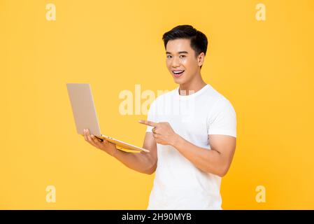 Souriant jeune homme asiatique beau pointant vers un ordinateur portable dans un studio isolé sur fond jaune Banque D'Images